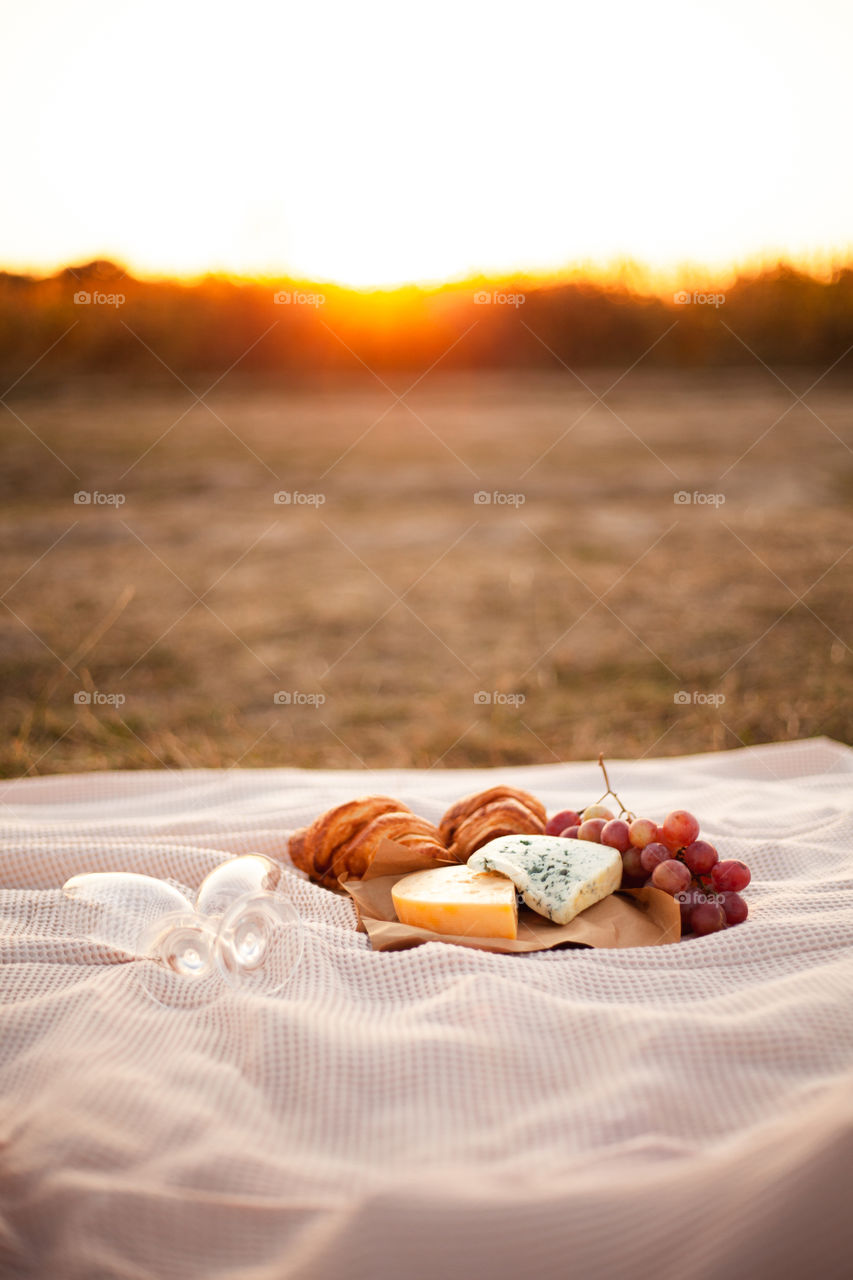 Romantic picnic for two on a sunset background. Two pieces of cheese, grapes, two croissants and two empty glasses lie on a blanket