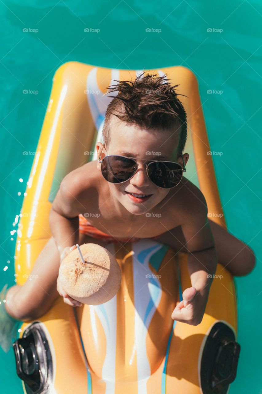 handsome boy of seven years with a coconut in his hands, sits on a swimming mattress, wearing glasses, spends time in the pool, smiles.  Summer mood