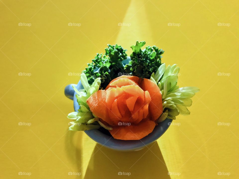 An overhead shot of a carrot rose in a little blue ceramic teacup framed with leaves fashioned from curly kale & dyed green daisy petals. Light pointed at the front of the cup creates an angled shadow behind the cup & through the teacup handle. 