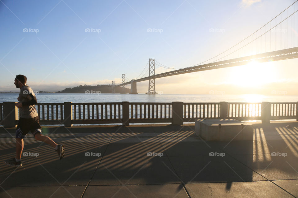 A jogger runs along a sunny wharf with the San Francisco Bay Bridge behind. 