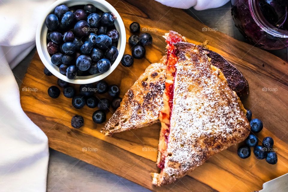 Peanut butter and blueberry jam french toast, served on a wooden board with a bowl of blueberries at the side.