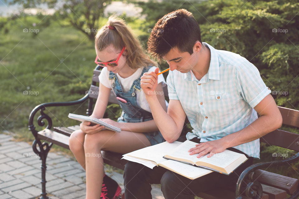 Students making the notes learning from books sitting on a bench in a park. Young boy wearing a blue shirt and dark jeans. Young blondie girl wearing jeans and sunglasses