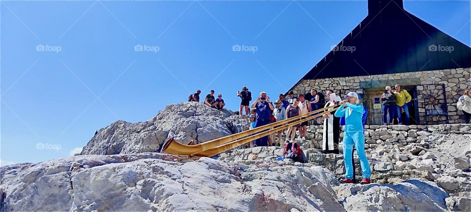 Massive “Alp” horns are blown by a couple of musicians in front of the “Mariä Heimsuchung”, “Visitation of Mary” chapel inaugurated by “Joseph Cardinal Ratzinger”, later “Pope Benedict XVI” in 1980. 2024. Hypnotic Productions