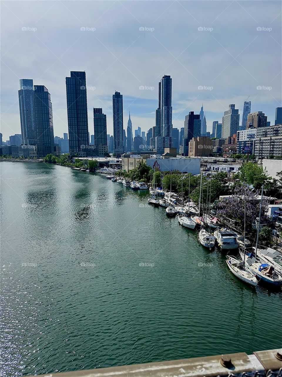 This is “Newtown Creek” with its many boats of various kinds seen from the “Pulaski Bridge” that connects LIC to “Greenpoint”, Bklyn. Across the “East River” in the distance we see “Manhattan” incl. the “Empire State Bldg”. 2024. Hypnotic Productions