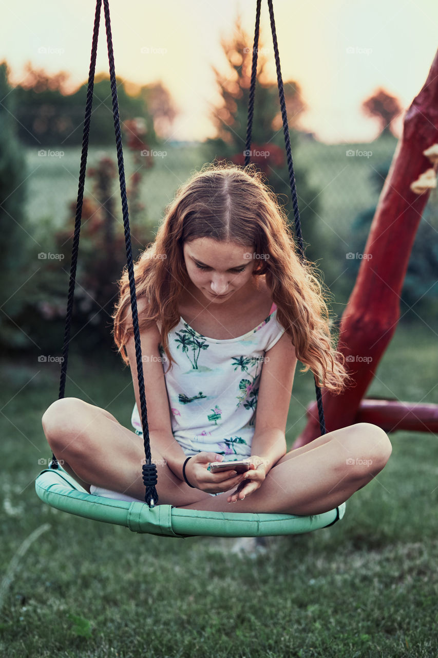Young woman using mobile phone smartphone sitting on swing in a backyard. Candid people, real moments, authentic situations