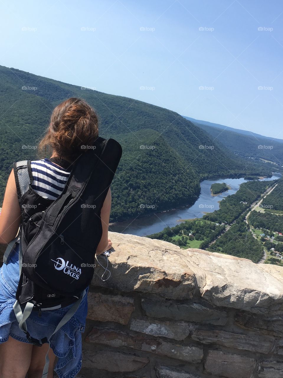 Girl with ukelele strapped to back contemplates river valley from above