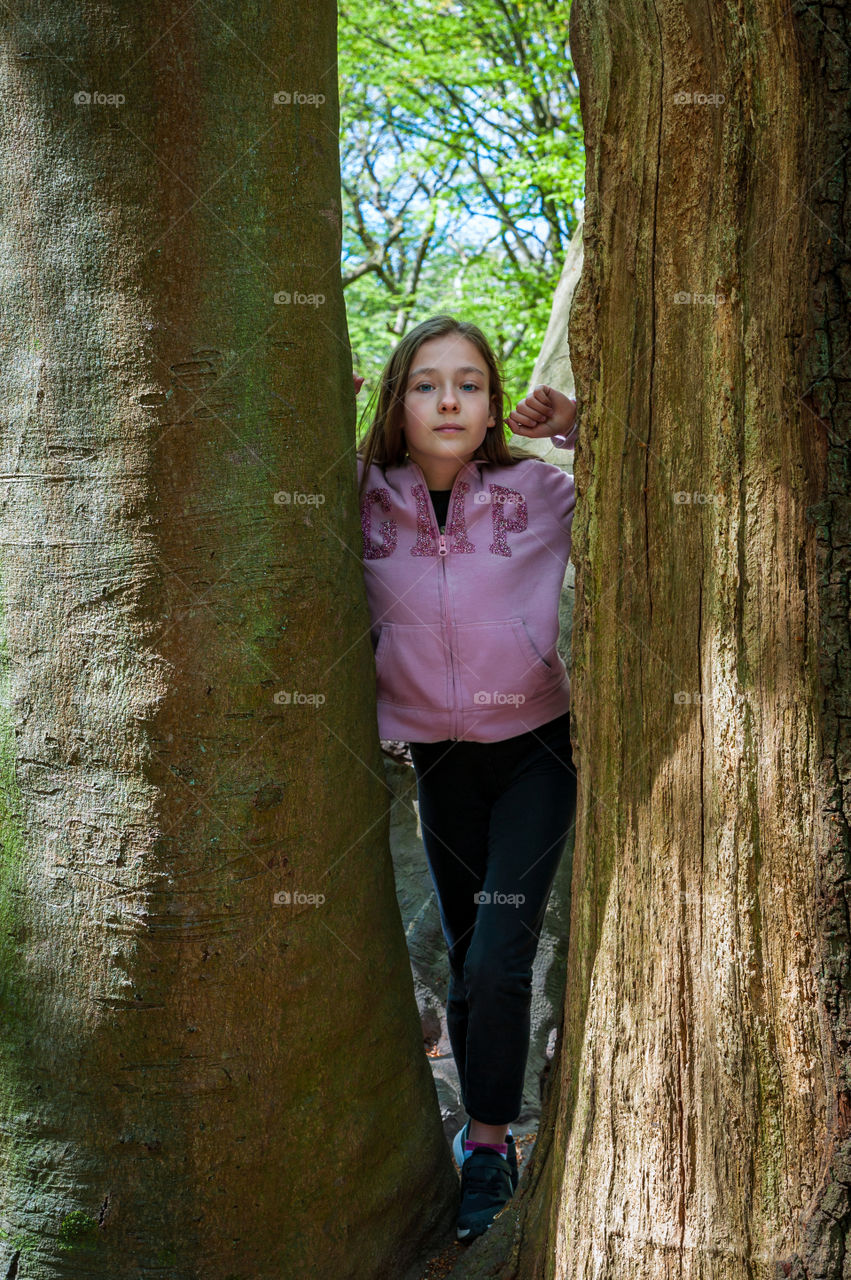 Young girl in pink zip hoodie standing between trees and looking into camera.