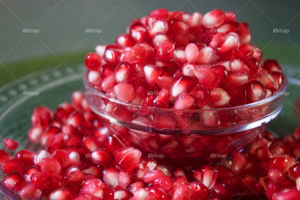 Closeup of fresh pomegranate seeds overflowing a small glass bowl against a green background