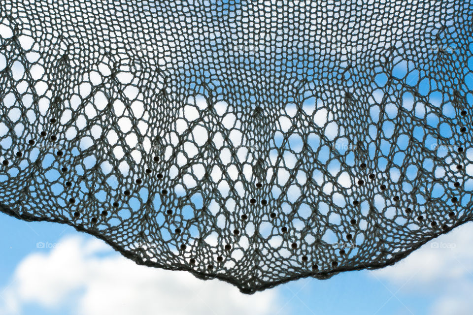 A lacy hand knit shawl draped in front of a blue sky spotted with white clouds