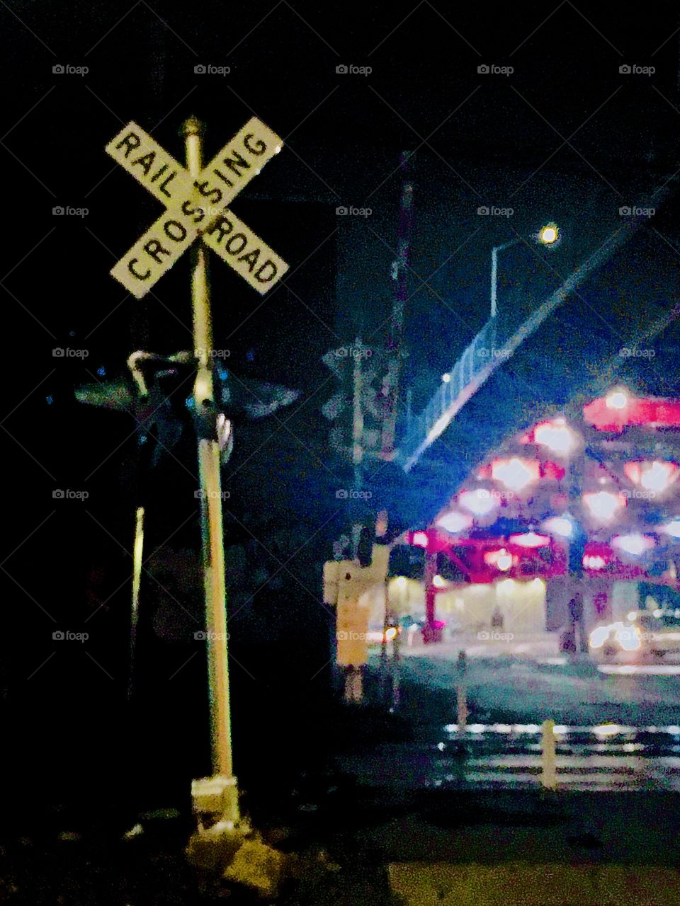 A crossed street sign warning of a “CROSSING” by the train tracks underneath the Pulaski Bridge late at night with an impressive multicolored neon light display in the background. Photo October 2020. Hypnotic Productions
