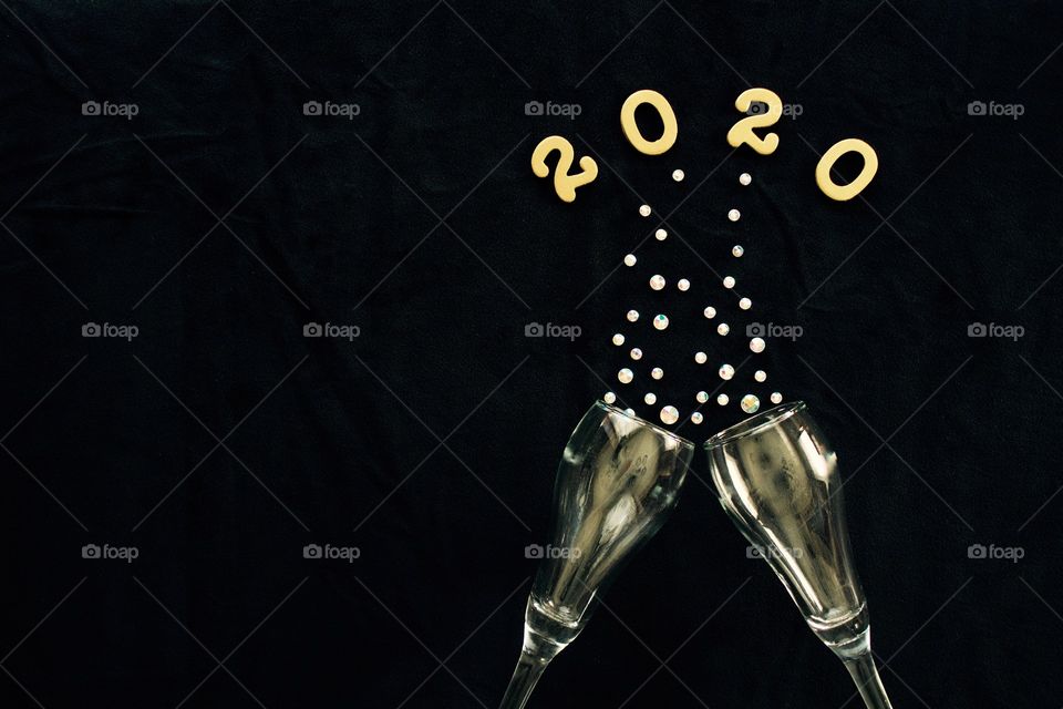 Flat lay of the numbers ‘2020’ in gold above gold-tinted champagne glasses with rhinestone bubbles on black background - copy space on left side of landscape format