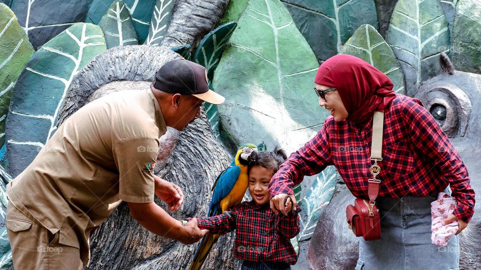 Batu, Indonesia - March 31, 2024 : Portrait of a little girl with her mother preparing to take a photo with a parrot on her shoulder at Batu Secret Zoo, assisted by zookeepers with happy expressions.
