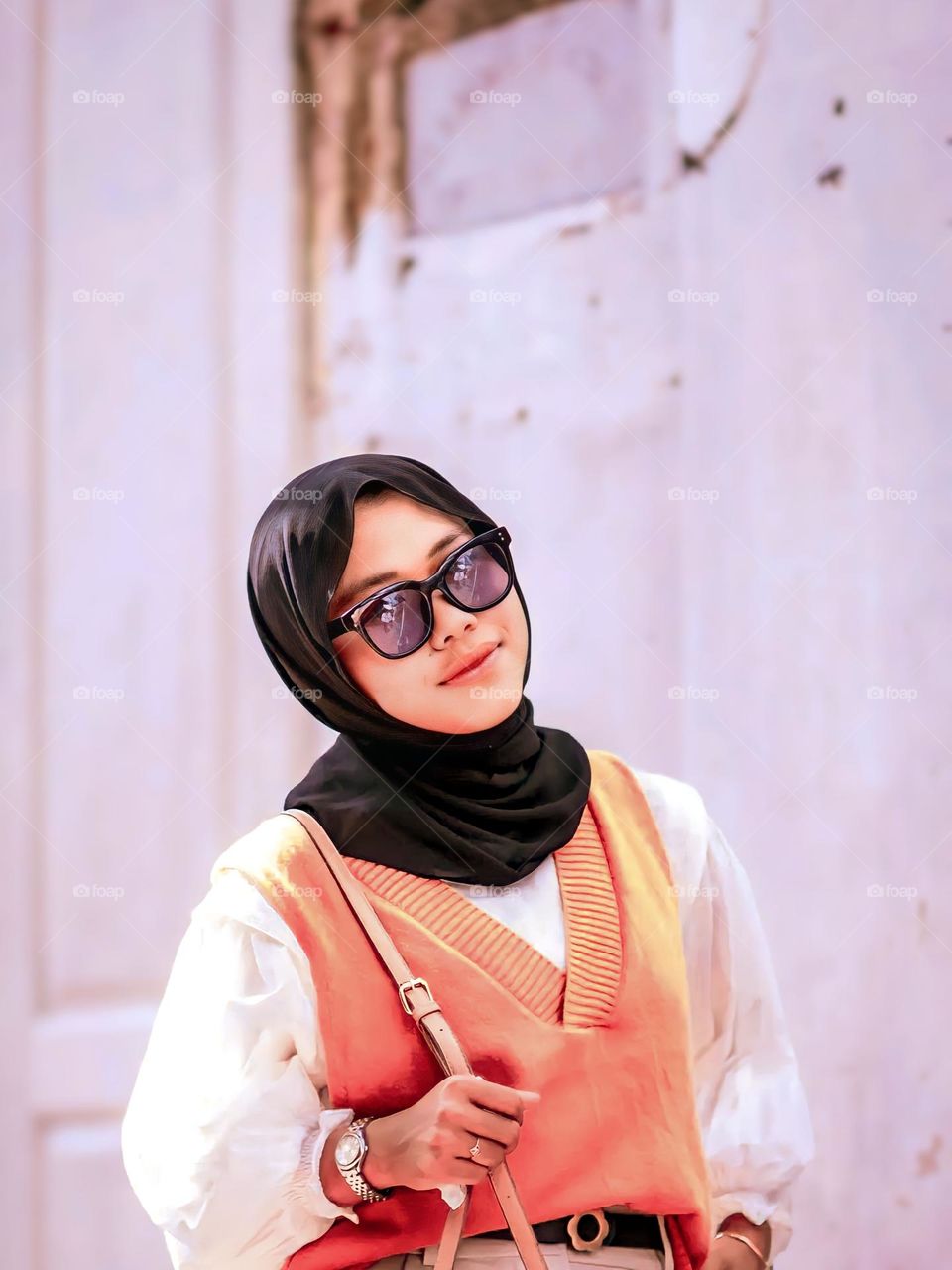 Close-up of young woman wearing black hijab and orange vest, standing in front of worn wall