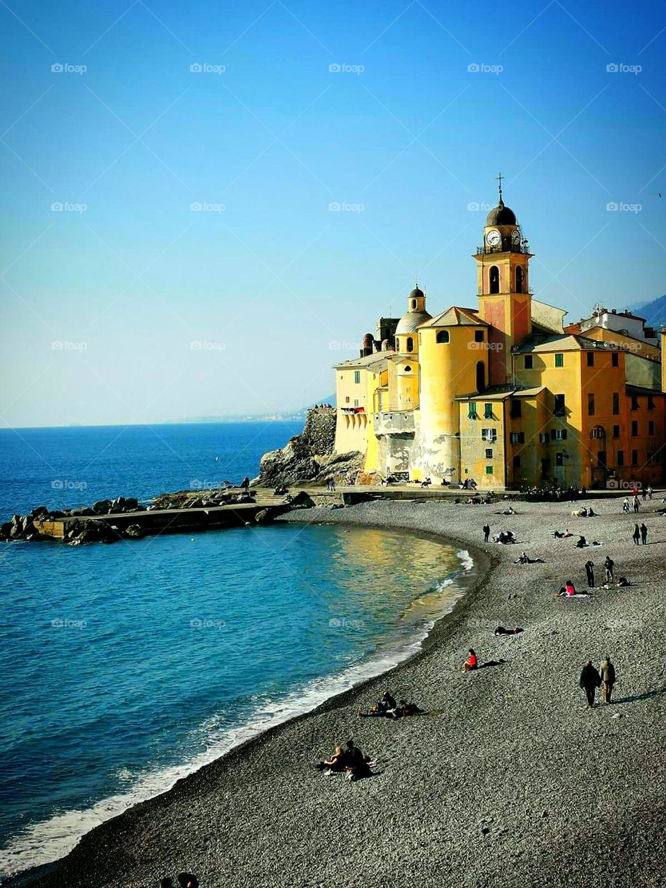 Camogli beach (Liguria). Italy. People sit and walk on the beach, basking in the first rays of the sun and looking at the sea. In the background is the Catholic Church a "Basilica di Santa"