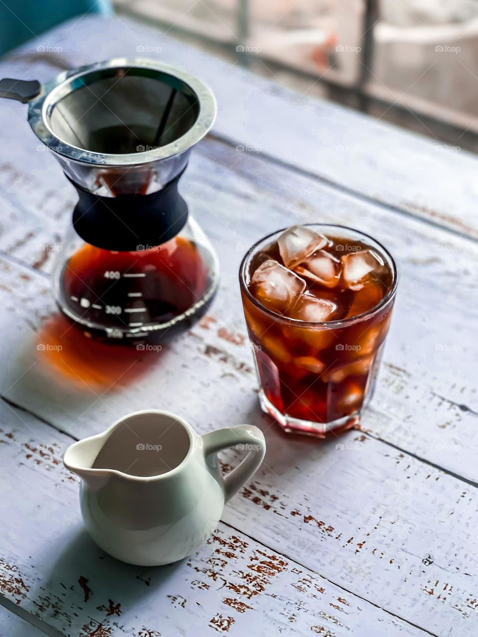 Iced coffee on a white wooden table with coffee dripper and ceramic milk jug on the side