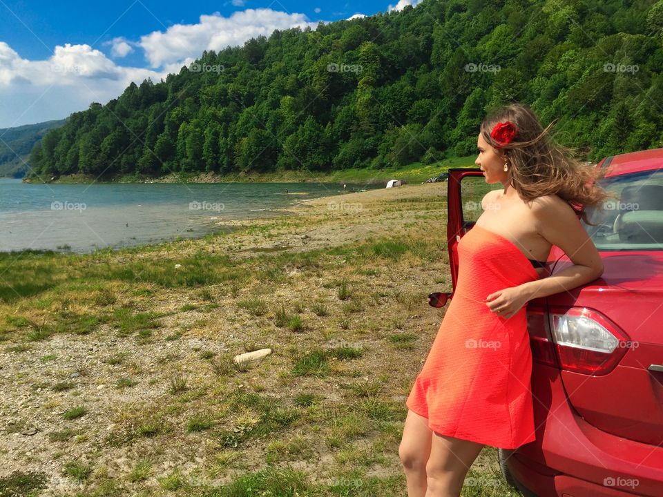 Woman in red dress standing near red car surrounded by lake and green forest in summer