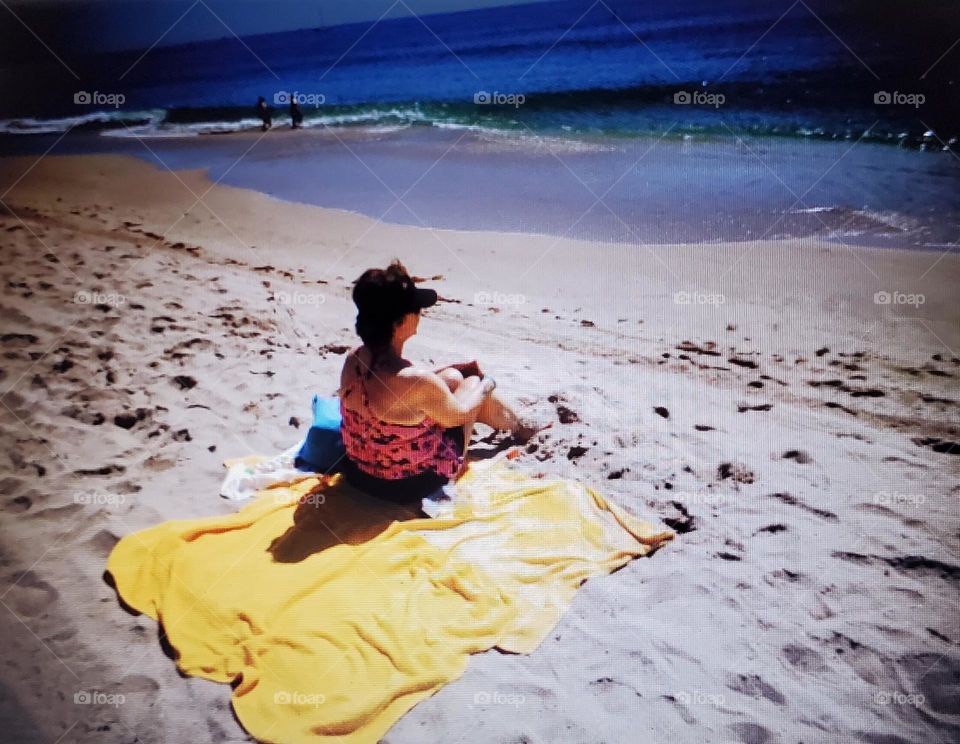 Woman or young girl sitting on a yellow blanket sun bathing at the beach. She's watching the waves, feet in sand, alone by herself. Sunny day at the beach enjoying sun wearing bathing suit & visor.