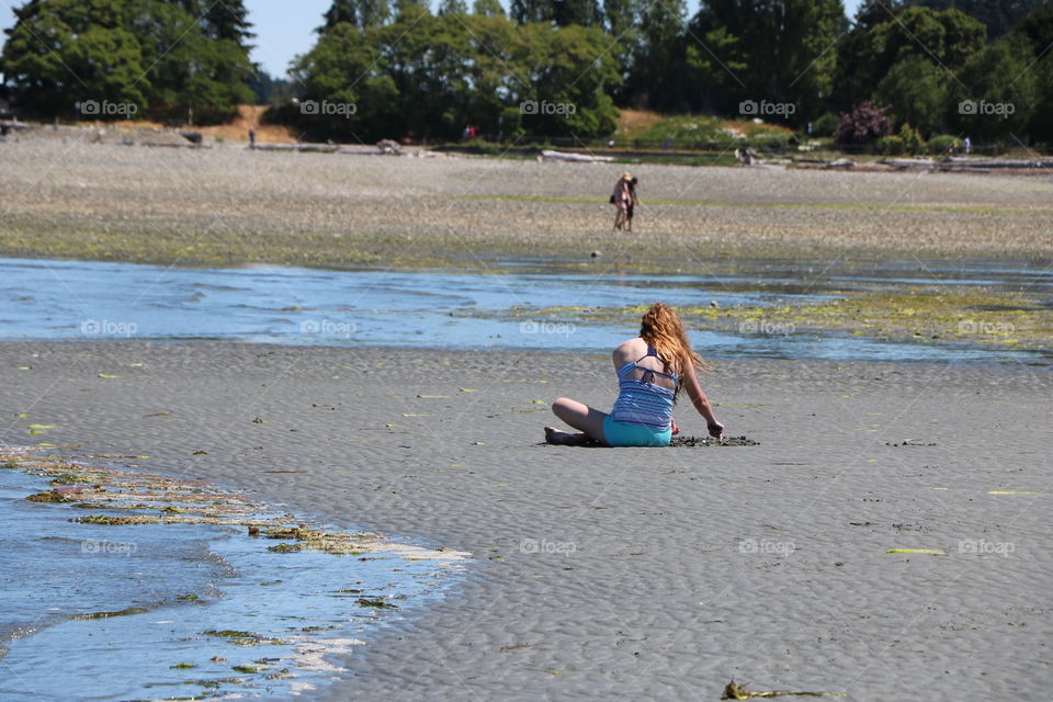 Wide sandy beach and a girl sitting and playing with the sand while tide coming and making little rivers of water all around