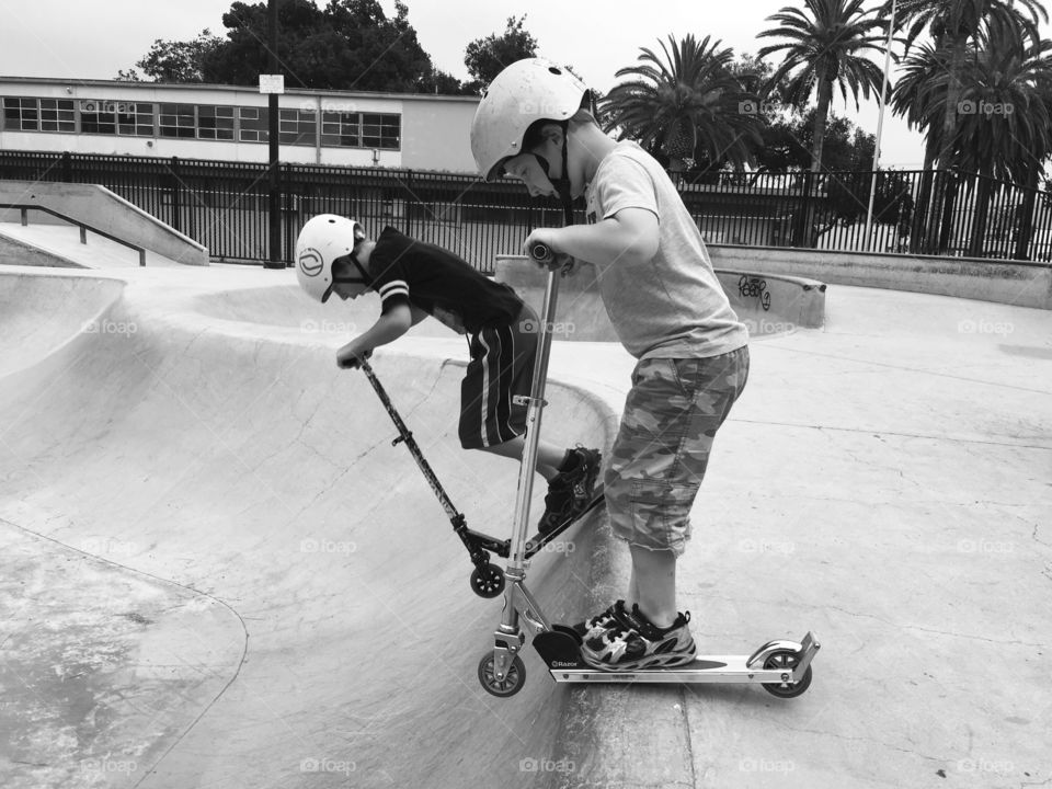 Learning how to go with the scooter on the abrupt slope in the skateboard park. The little brother learns from his older brother the skill and the courage.