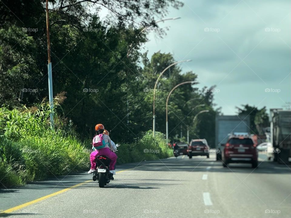 Man and woman riding a motorcycle in the freeway with trees and cars around them.