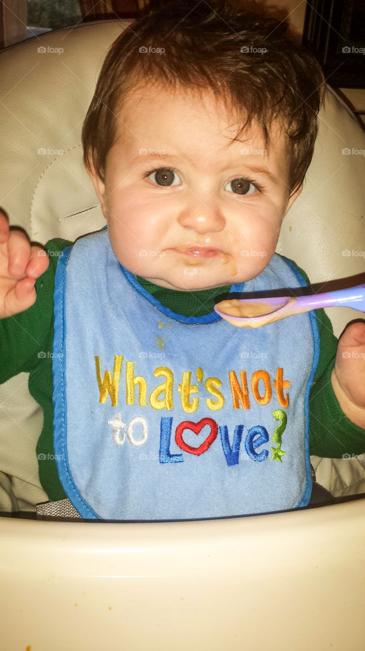 A baby boy wearing a "What's Not to Love?" bib is clearly not loving anything about his baby food!