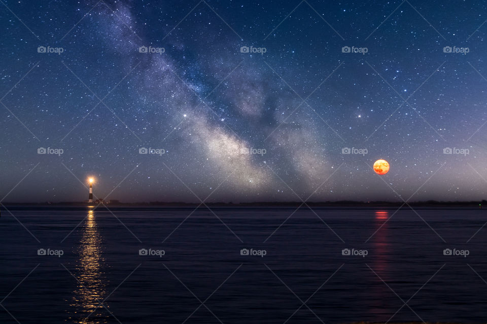 The Milky Way rising in the night sky, with a lighthouse and full moon brightly reflecting in a body of water. 