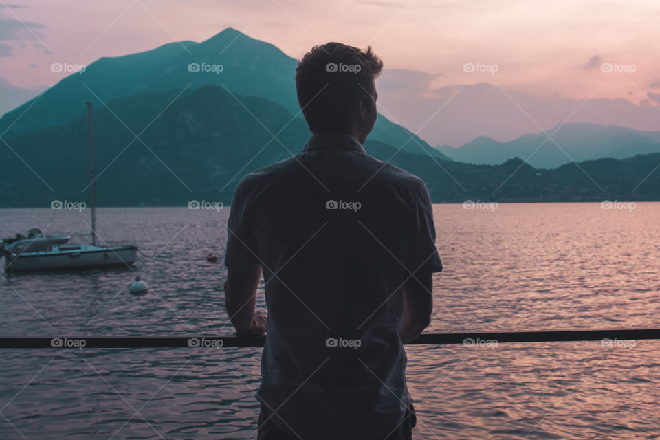 A man looks out into the sun setting over the mountains of lake como in Italy as boats dock for the evening