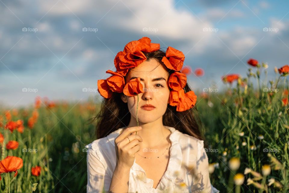 Beautiful woman holding a poppy flower in front of her face, while being surrounded by a field of poppy flowers.