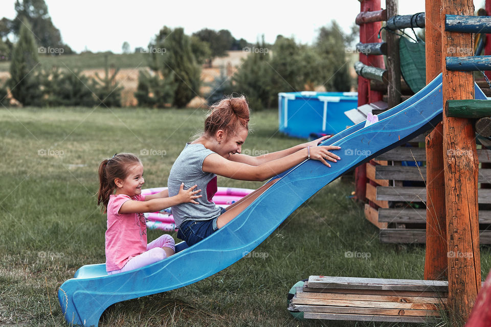 Teenage girl playing with her younger sister in a home playground in a backyard. Happy smiling sisters having fun on a slide together on summer day. Real people, authentic situations