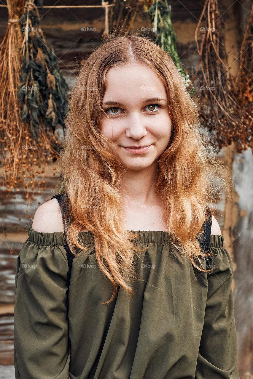 Portrait of young woman  standing in front of old cottage and hung some withered species of herbs. Girl is wearing green dress, looking at camera