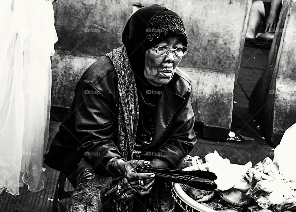 An old woman is hawking on the side of the street, which is selling homemade traditional food