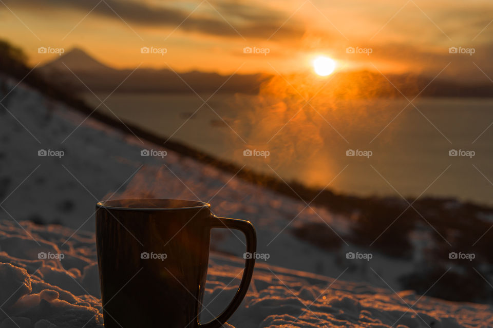 In the snow there is a mug with hot coffee from which steam rises and through it you can see the sun setting behind the mountains