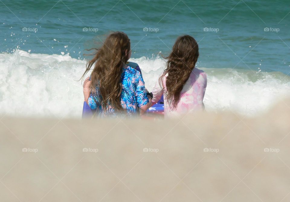 Two young girls children at the beach on vacation facing a wave from the Atlantic Ocean in front of a seashore sand hill with body boards and long hair with modest blue and pink swimsuits.