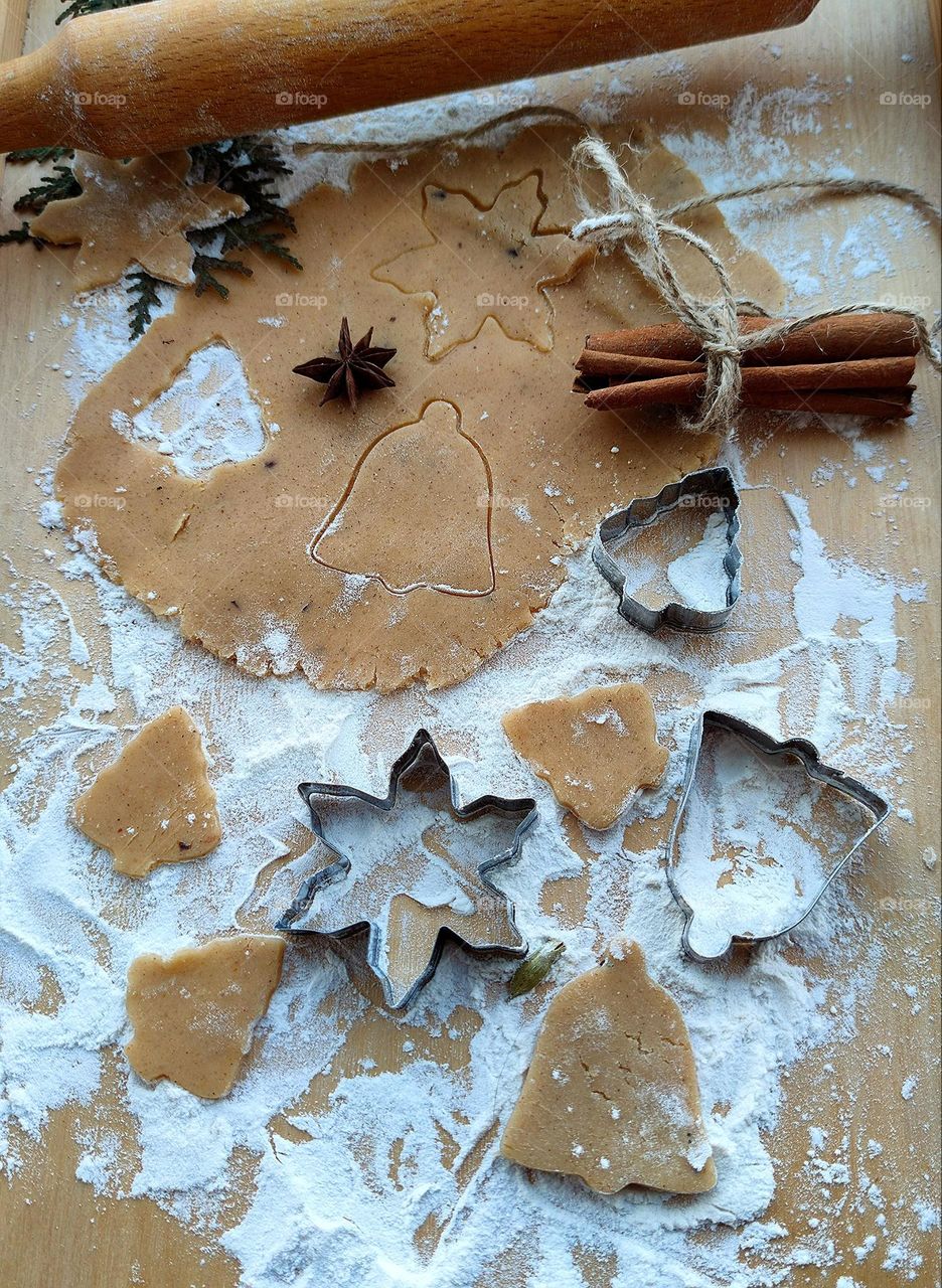 On a board sprinkled with white flour lies a rolled out dough with squeezed out figures. Nearby are figures of their dough, metal molds, a stack of cinnamon sticks, which is tied with twine. In the background lies a wooden rolling pin.