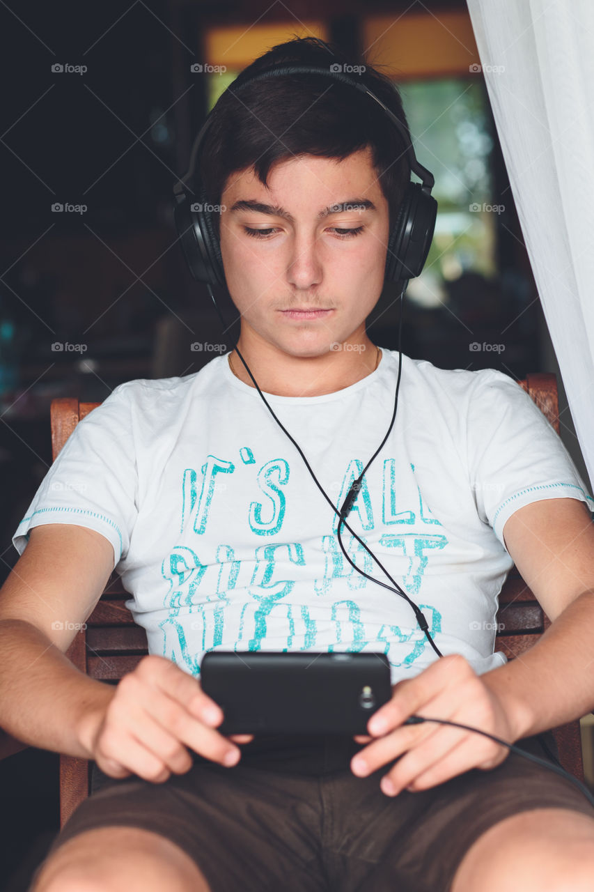 Concentrated on mobile phone. Young boy sitting at the patio door and listening to music from mobile phone