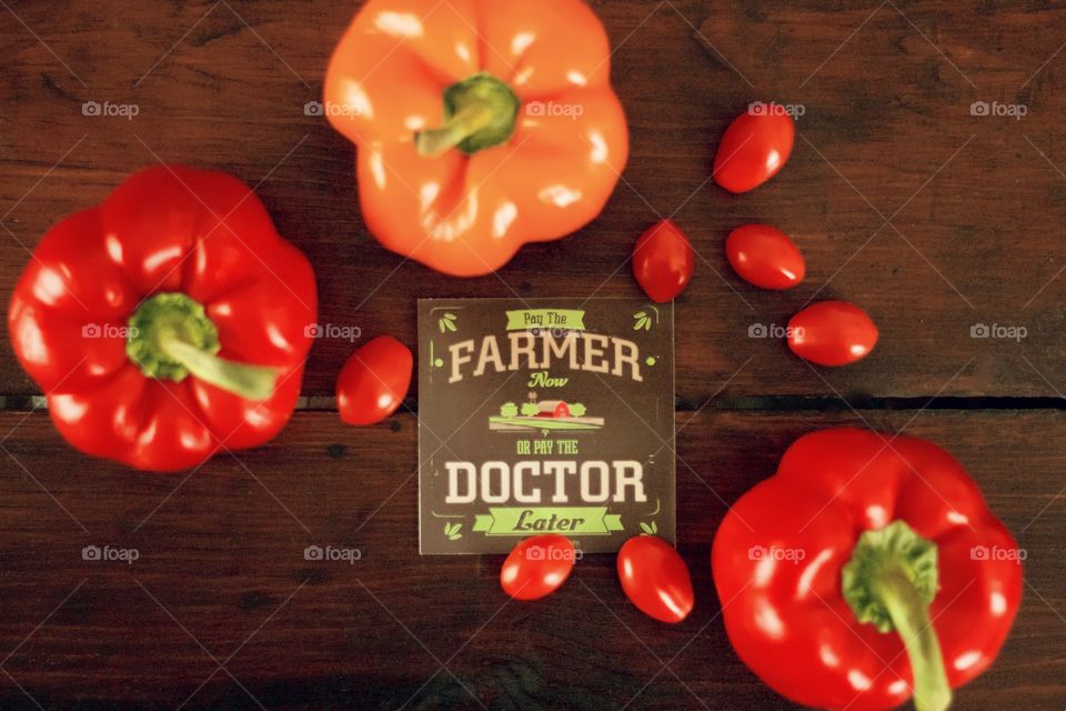 Flat lay of “Pay the Farmer Now or Pay the Doctor Later” sign with red and orange bell peppers and grape tomatoes on dark wooden surface