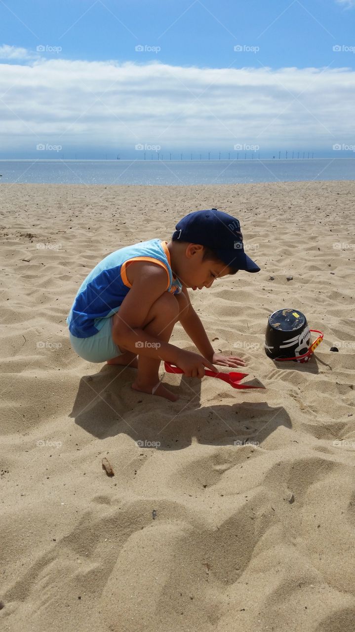 small kid playing on the sandy beach with a spade in a hot summer day wearing blue hat and top