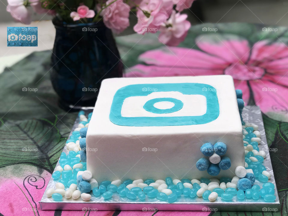 A foap wedding cake with the logo made of white & turquoise fondant. Daisies of turquoise gummies & marshmallows on the side of the cake & turquoise & white jelly beans on the cake base. All on a green & pink table cloth with a vase of pink roses. ๐
