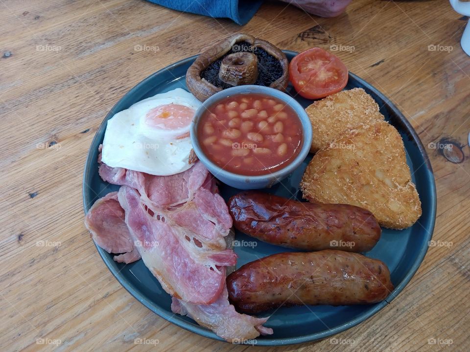 A large Full English Breakfast of Bacon, Two Sausages, Fried Egg, Baked Beans in a small Bowl, Two Hash Browns, Grilled Tomato and Mushroom, served in a Blue Plate on a Wooden Table.