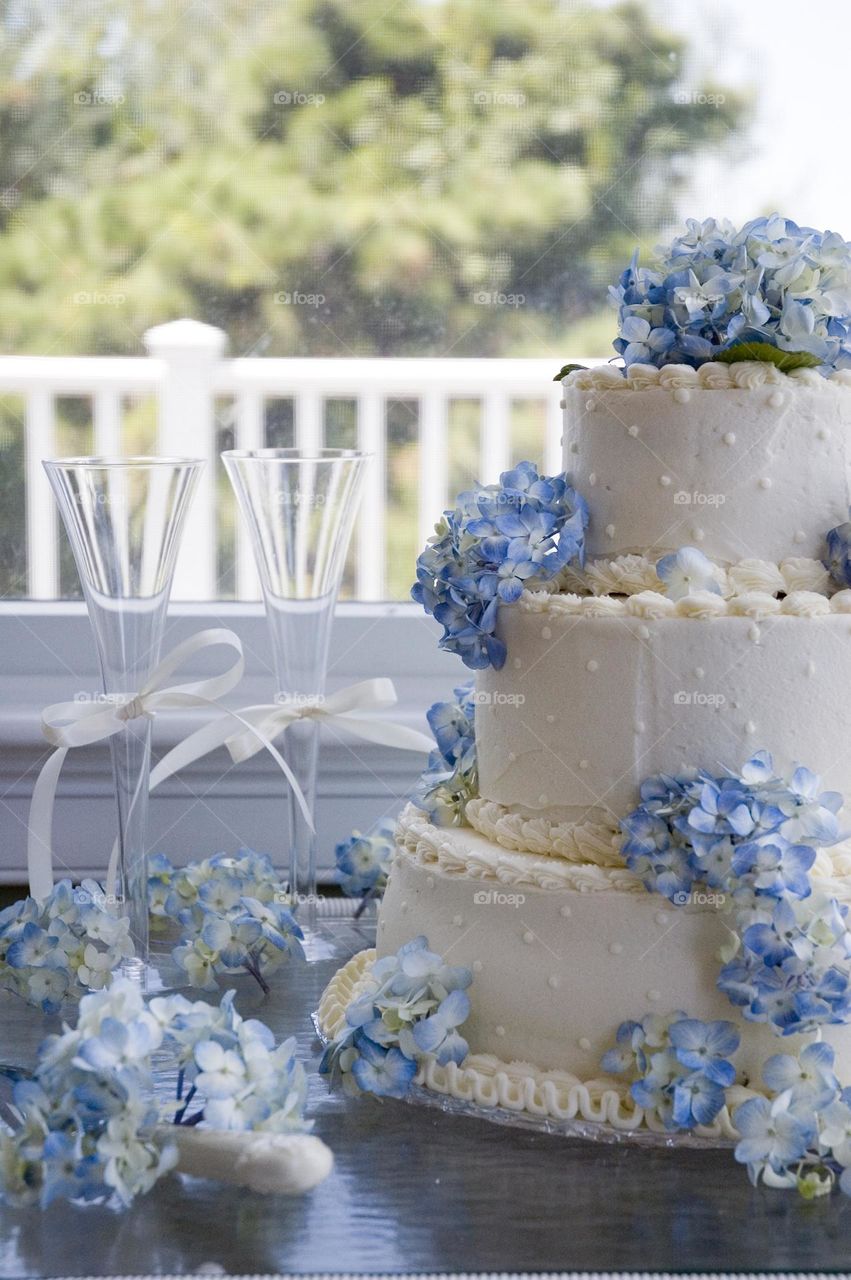 A still life of a three tier wedding cake with light blue hydrangeas and two champagne flutes with white ribbons 