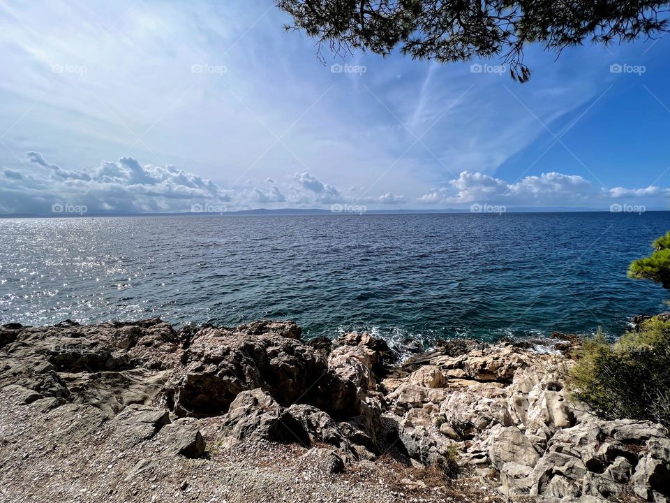 Sunny day landscape horizon with the low clouds on the blue sky over the turquoise sea water surface and rocky shore coastline