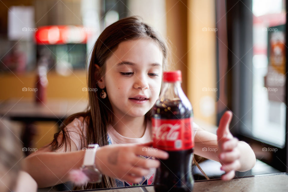 Cute, little brunette girl sitting in a bright restaurant waiting for her pizza and excited about drinking her favorite drink, Coke. 
