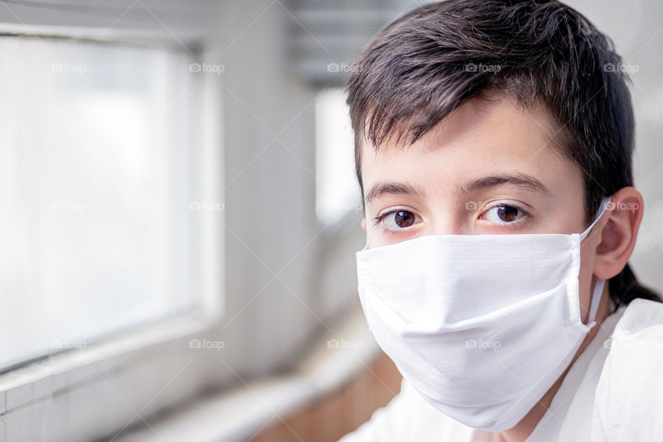 Portrait of child in white protective mask near the window. Concept of 2020.