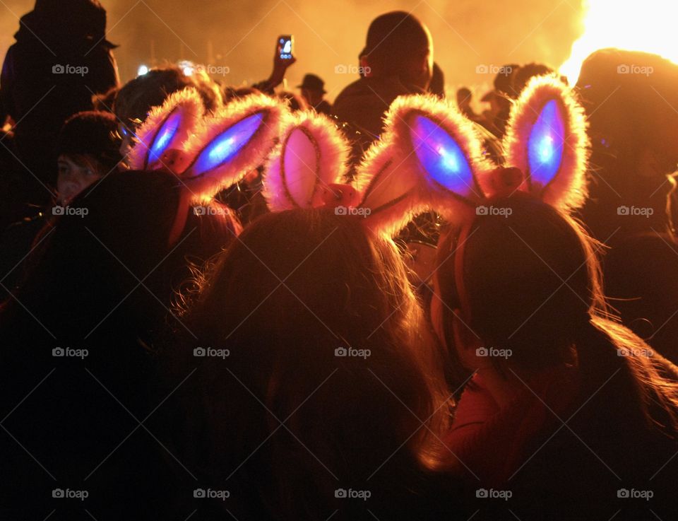 3 teenagers wearing light up bunny ears stand among the crowd watching a bonfire, the air is orange and smokey 