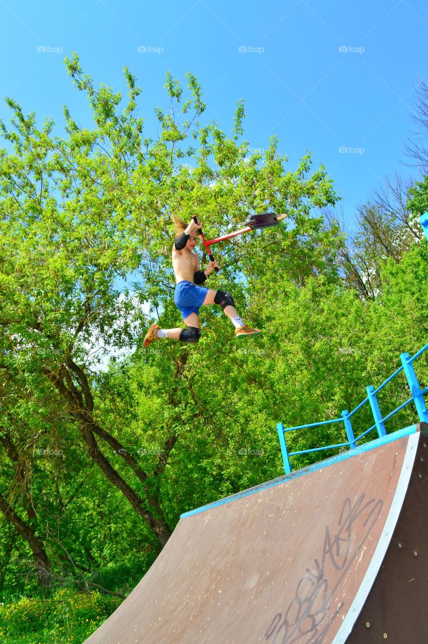 guy athlete performs a trick in the air on a scooter. in the frame a man flying over the ramp trees and blue sky.