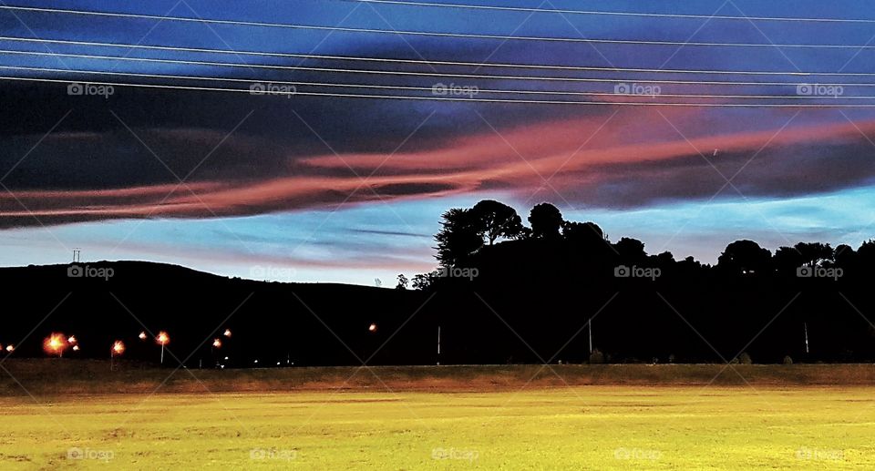 As The sun sets at Napier the lights from the hockey field lights up the grass. the hills and trees dark the colors of the last suns rays vibrant in the heavens powerlines overhead.