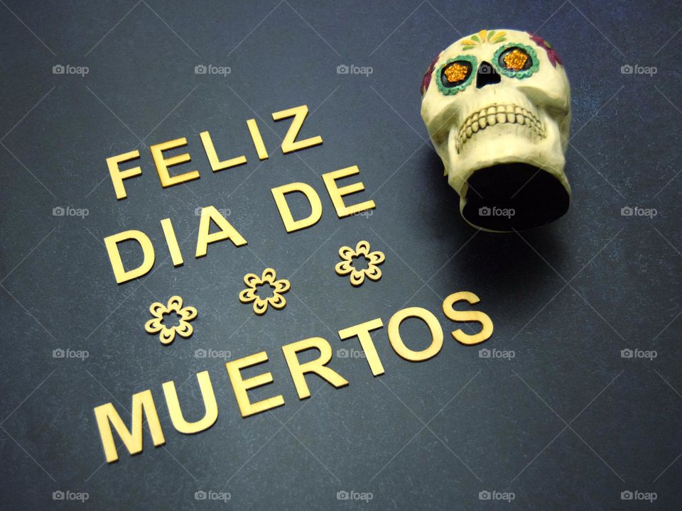 A simple flatlay featuring wooden letters arranged to form the phrase “Feliz Dia De Los Muertos” styled alongside simple but beautiful wooden flowers & a traditional & beautifully coloured “Calavera” or “sugar skull”