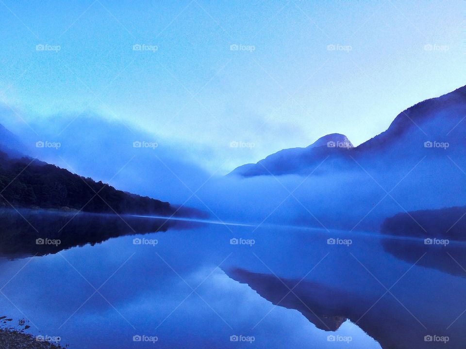 Fog settles over a glass lake that reflects the landscape and mountains in Fiordland National Park in South Island, New Zealand.  