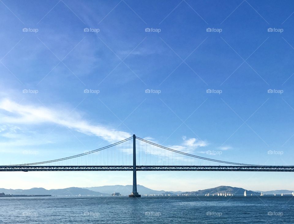 Bay Bridge over Pacific Ocean full of sailboats against a backdrop of faded mountains. 