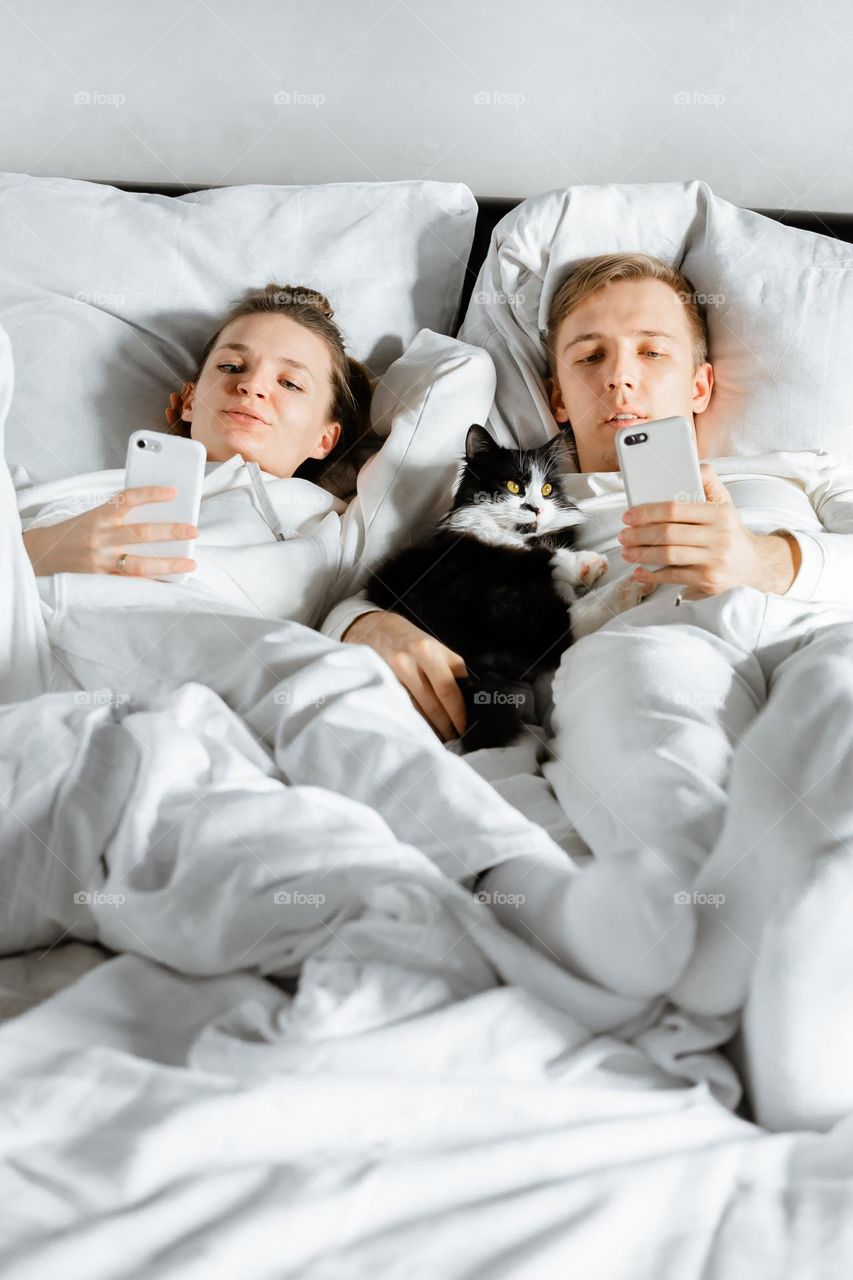 Couple using phone chatting in social media laying on white bed with black cat 